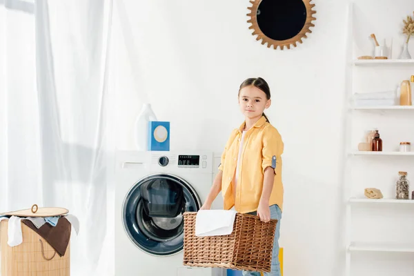 Kind in der Nähe der Waschmaschine hält Korb mit Handtuch in der Waschküche — Stockfoto
