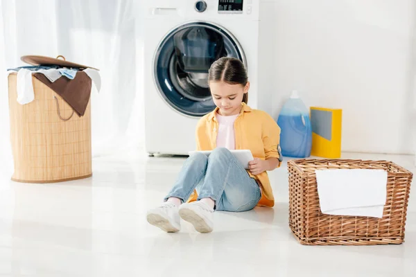 Enfant en chemise jaune et jeans assis avec tablette numérique dans la buanderie — Photo de stock