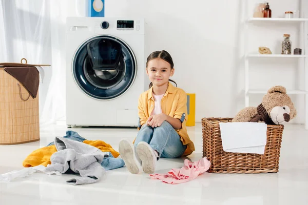 Bambino in camicia gialla e jeans seduto vicino a vestiti e cestini in lavanderia — Foto stock