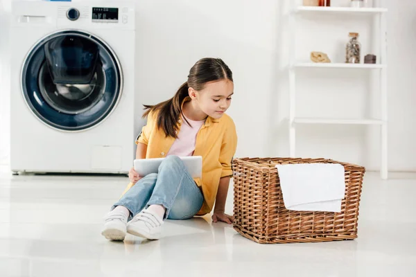 Kind in gelbem Hemd und Jeans sitzt mit digitalem Tablet und sucht Korb in Waschküche — Stockfoto