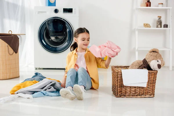 Child in yellow shirt and jeans sitting and putting clothes to basket in laundry room — Stock Photo