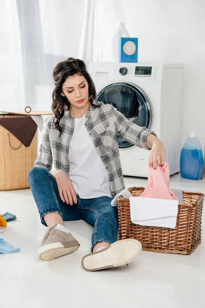 Atractiva mujer sentada en el suelo y poniendo ropa a la cesta en el cuarto de lavado - foto de stock