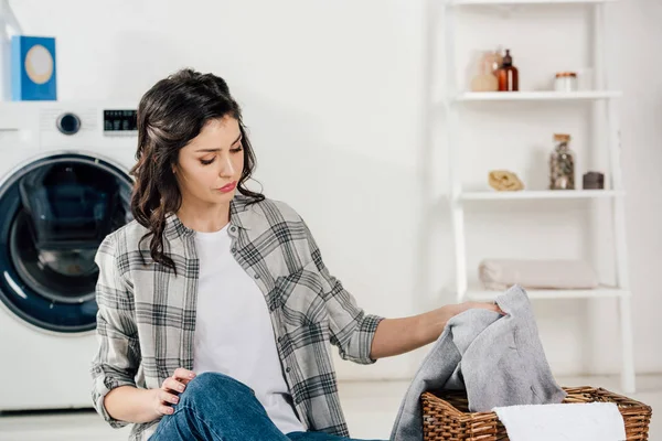 Selective focus of attractive woman sitting on floor and putting clothes to basket in laundry room — Stock Photo