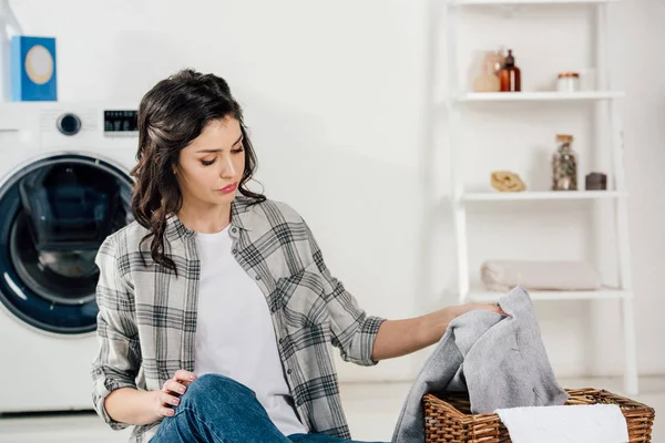 Foyer sélectif de femme attrayante assise sur le sol et mettre des vêtements au panier dans la buanderie — Photo de stock