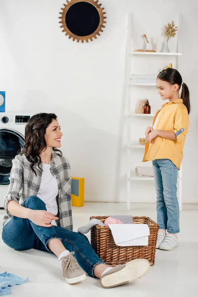 Madre sentada en el suelo cerca de la cesta y mirando a su hija en camisa amarilla en el cuarto de lavado - foto de stock