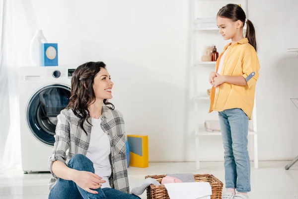 Mother sitting on floor near basket, looking to daughter in yellow shirt and smiling in laundry room — Stock Photo