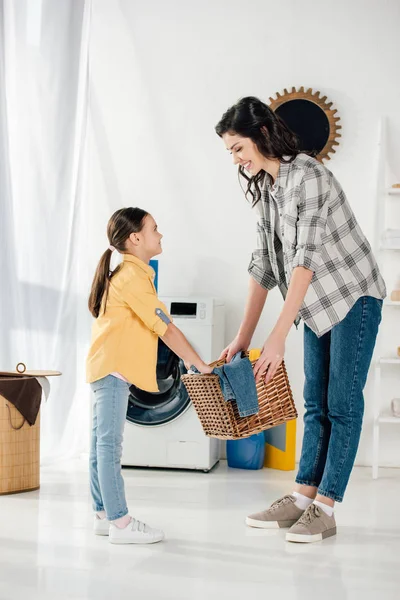 Hija en camisa amarilla y madre en camisa gris sosteniendo cesta y sonriendo en la lavandería - foto de stock