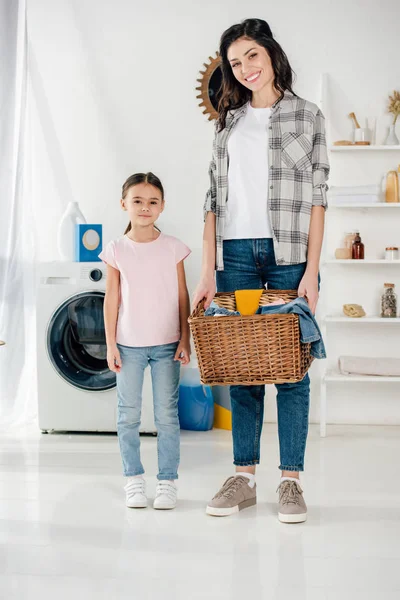Hija en camiseta rosa de pie cerca de la madre sonriente en camisa gris con cesta y en el cuarto de lavado - foto de stock