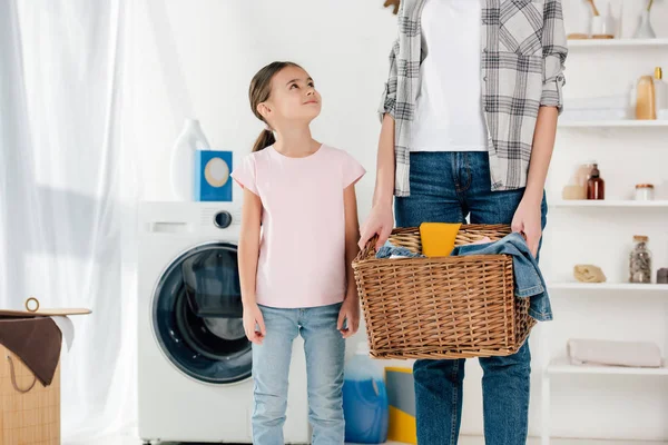 Hija en camiseta rosa mirando a la madre en camisa gris con cesta en lavadero - foto de stock
