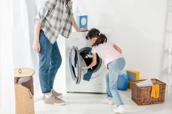 Filha em camiseta rosa colocando roupas na máquina de lavar wile mãe de pé na lavanderia — Fotografia de Stock