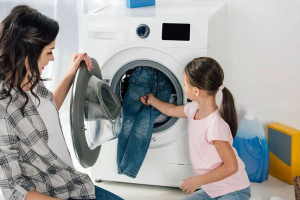 Filha em camiseta rosa e mãe colocando roupas na máquina de lavar roupa na lavanderia — Fotografia de Stock