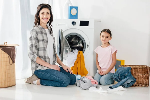 Hija en camiseta rosa y madre en camisa gris sentada en el suelo cerca de ropa dispersa y lavadora en la sala de lavandería - foto de stock