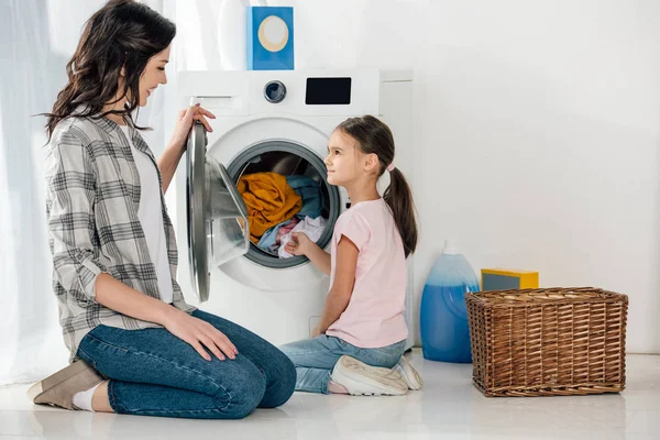 Filha em camiseta rosa e mãe em camisa cinza sentado no chão perto da máquina de lavar roupa com roupas na lavanderia — Fotografia de Stock