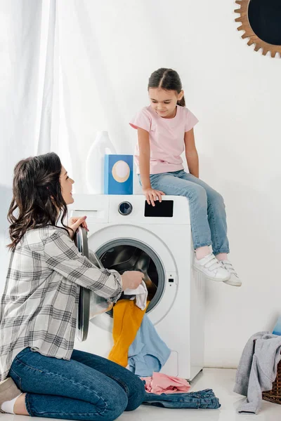 Figlia in t-shirt rosa seduta sulla lavatrice madre astuta in camicia grigia che mette i vestiti in lavanderia — Foto stock