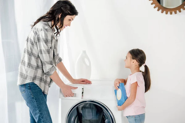 Hija en rosa camiseta sosteniendo lavado en polvo madre astuta en gris camisa apertura lavadora en la sala de lavandería - foto de stock