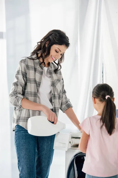 Mère en chemise grise versant du détergent à lessive liquide dans la laveuse avec fille debout dans la buanderie — Photo de stock