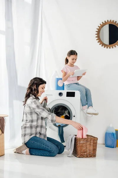 Hija en camiseta sentada en la lavadora con tableta digital madre astuta en camisa gris tomando ropa de la cesta en la sala de lavandería - foto de stock