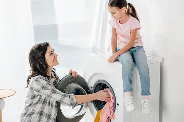 Figlia in t-shirt rosa seduta sulla lavatrice madre astuta in camicia grigia che mette i vestiti in lavanderia — Foto stock