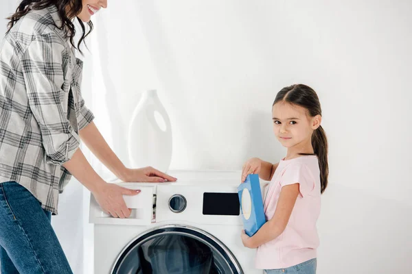 Daughter in pink t-shirt holding washing powder wile mother opening washer in laundry room — Stock Photo