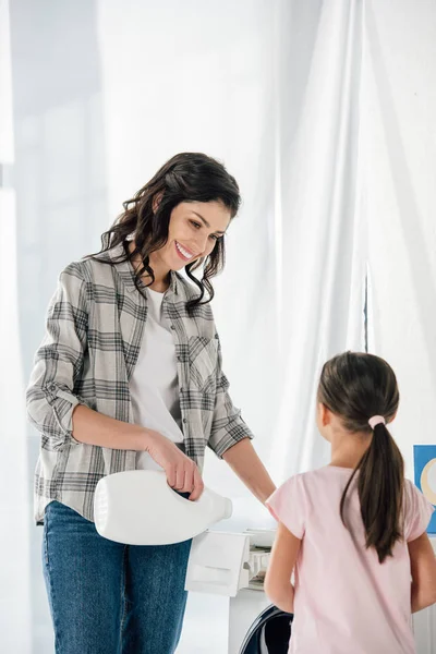 Madre in camicia grigia versando detersivo per bucato liquido in lavatrice figlia astuta in t-shirt rosa in piedi vicino in lavanderia — Foto stock