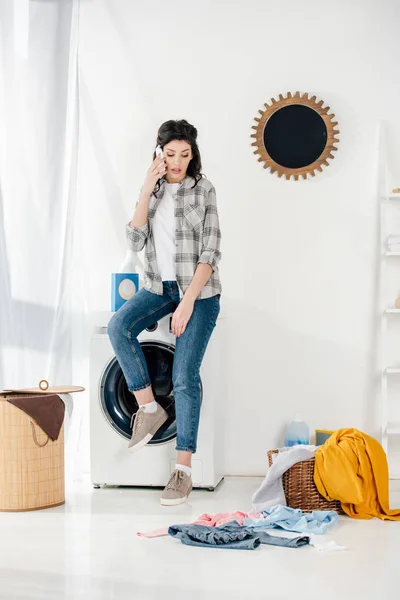 Mujer sentada en la lavadora y hablando por teléfono inteligente en la sala de lavandería - foto de stock