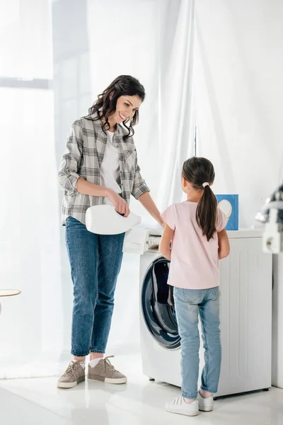 Madre verter detergente líquido en lavadora hija astuta en camiseta rosa de pie cerca en la sala de lavandería - foto de stock