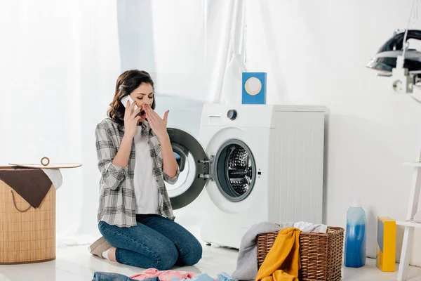 Mujer sentada cerca de la lavadora, hablando por teléfono inteligente y bostezando en la sala de lavandería - foto de stock