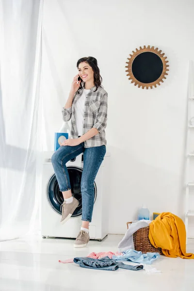 Mujer en camisa gris y jeans sentados en la lavadora y hablando en el teléfono inteligente en la sala de lavandería - foto de stock