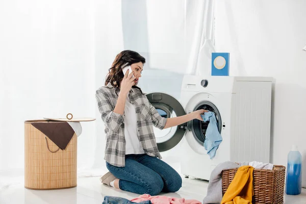 Mujer poniendo la ropa en la lavadora y hablando en el teléfono inteligente en la sala de lavandería - foto de stock
