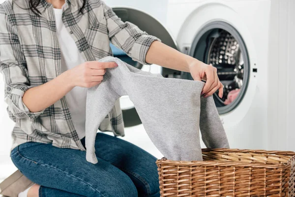 Vista recortada de la mujer sosteniendo la ropa cerca de la lavadora en la sala de lavandería - foto de stock