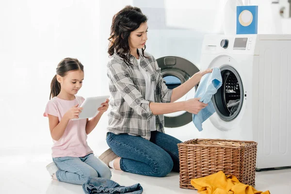 Mutter hält Kleidung in der Nähe in der Waschmaschine, Tochter in rosa T-Shirt sitzt mit digitalem Tablet in Waschküche — Stockfoto