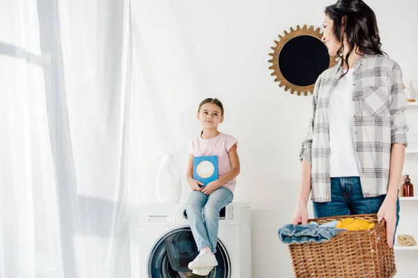 Foco seletivo da filha sentada na máquina de lavar com lavagem em pó wile mãe segurando cesta na lavanderia — Fotografia de Stock