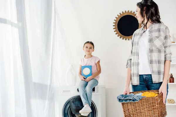 Foyer sélectif de la fille assise sur la rondelle avec la poudre à laver wile mère tenant panier dans la buanderie — Photo de stock