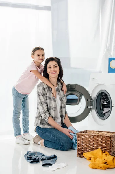 Filha abraçando mãe perto lavadora e cesta na lavanderia — Fotografia de Stock