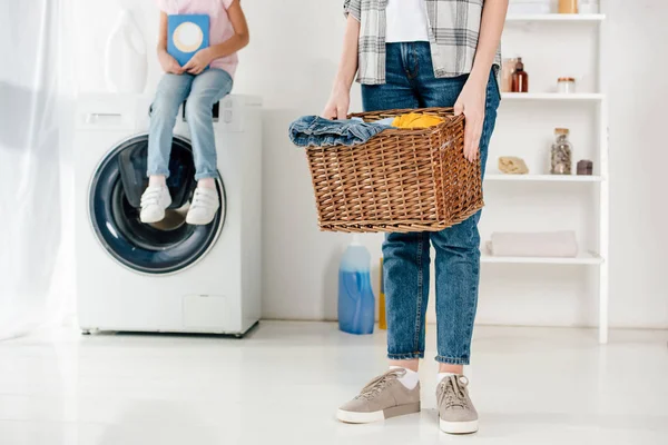 Обрізаний вид на дочку, що сидить на пральній машині з порошком, мати тримає кошик у пральні — стокове фото