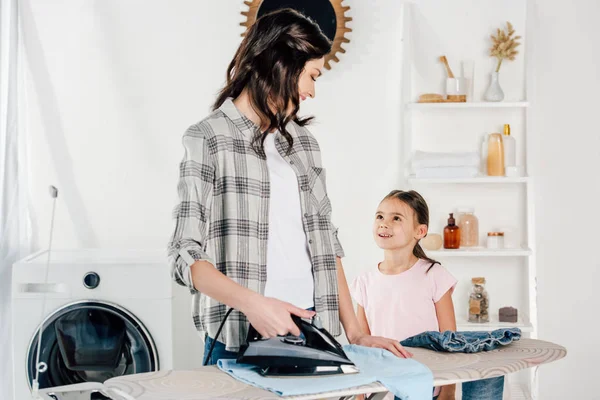 Madre planchado y mirando a la hija de pie cerca en la sala de lavandería - foto de stock