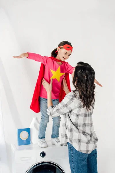 Mãe em camisa cinza tocando filha em terno caseiro vermelho com sinal de estrela na máquina de lavar roupa na lavanderia — Fotografia de Stock
