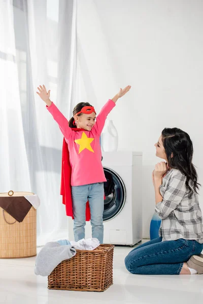Mother in grey shirt sitting on floor near daughter in red homemade suit with star sign in laundry room — Stock Photo