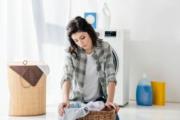 Selective focus of upset woman in grey shirt sitting near basket in laundry room — Stock Photo