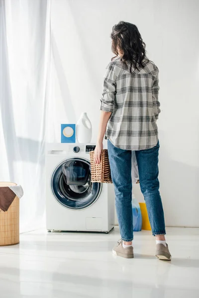 Visão traseira da mulher em camisa cinza e jeans segurando cesta perto lavadora na lavanderia — Fotografia de Stock
