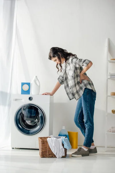 Woman in grey shirt and jeans standing near washer woman and holding backache in laundry room — Stock Photo