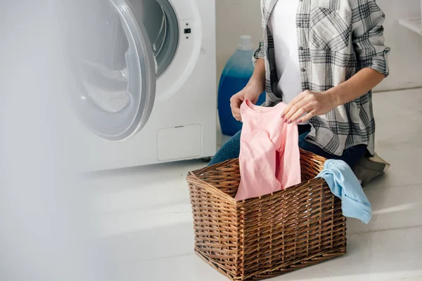 Vue recadrée de la femme en chemise grise et jeans mettant des vêtements dans le panier près de la laveuse dans la buanderie — Photo de stock