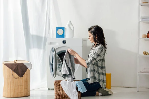 Mujer en camisa gris y jeans poniendo ropa en la cesta cerca de la lavadora en la sala de lavandería - foto de stock