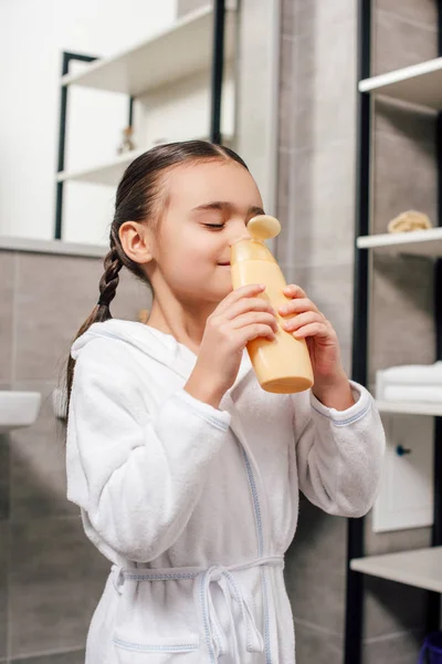 Child in white bathrobe sniffing shower gel in bathroom — Stock Photo