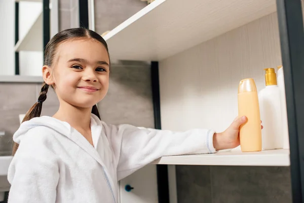 Cute child in white bathrobe taking shower gel from shelf in bathroom — Stock Photo