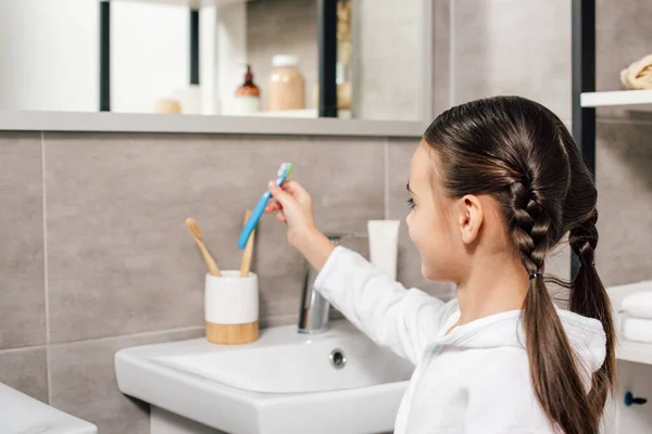 Милый ребенок в белом халате принимает зубную пасту в ванной комнате — стоковое фото