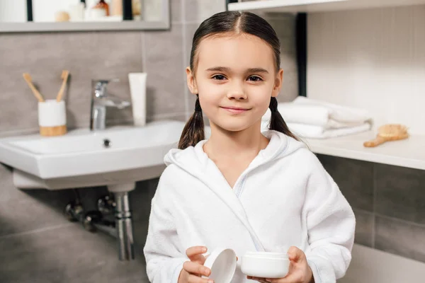 Мила дитина в білому халаті тримає косметичний крем у ванній — стокове фото