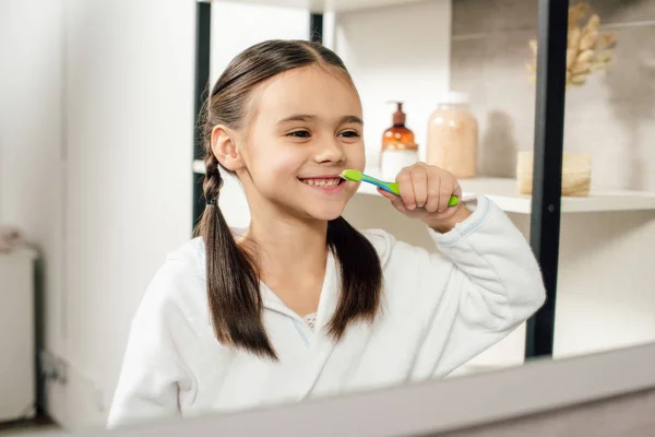 Selective focus of child in white bathrobe looking to mirror and brushing teeth in bathroom — Stock Photo