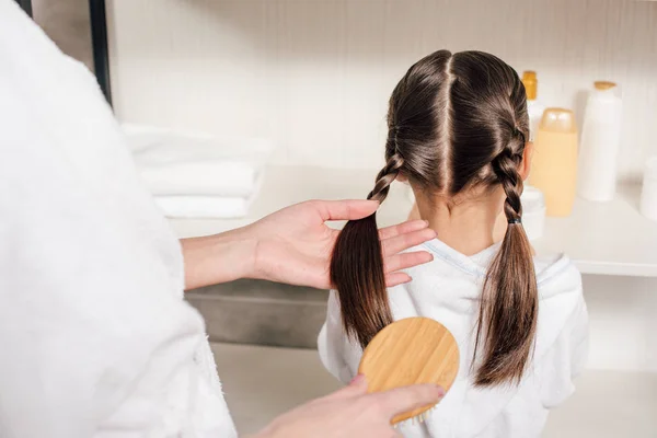 Mother in white bathrobe combing daughter hair in bathroom — Stock Photo