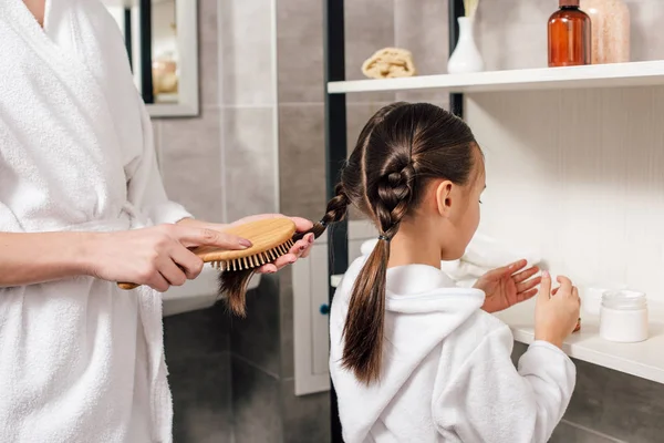 Madre in accappatoio bianco pettinatura figlia capelli vicino scaffali in bagno — Foto stock